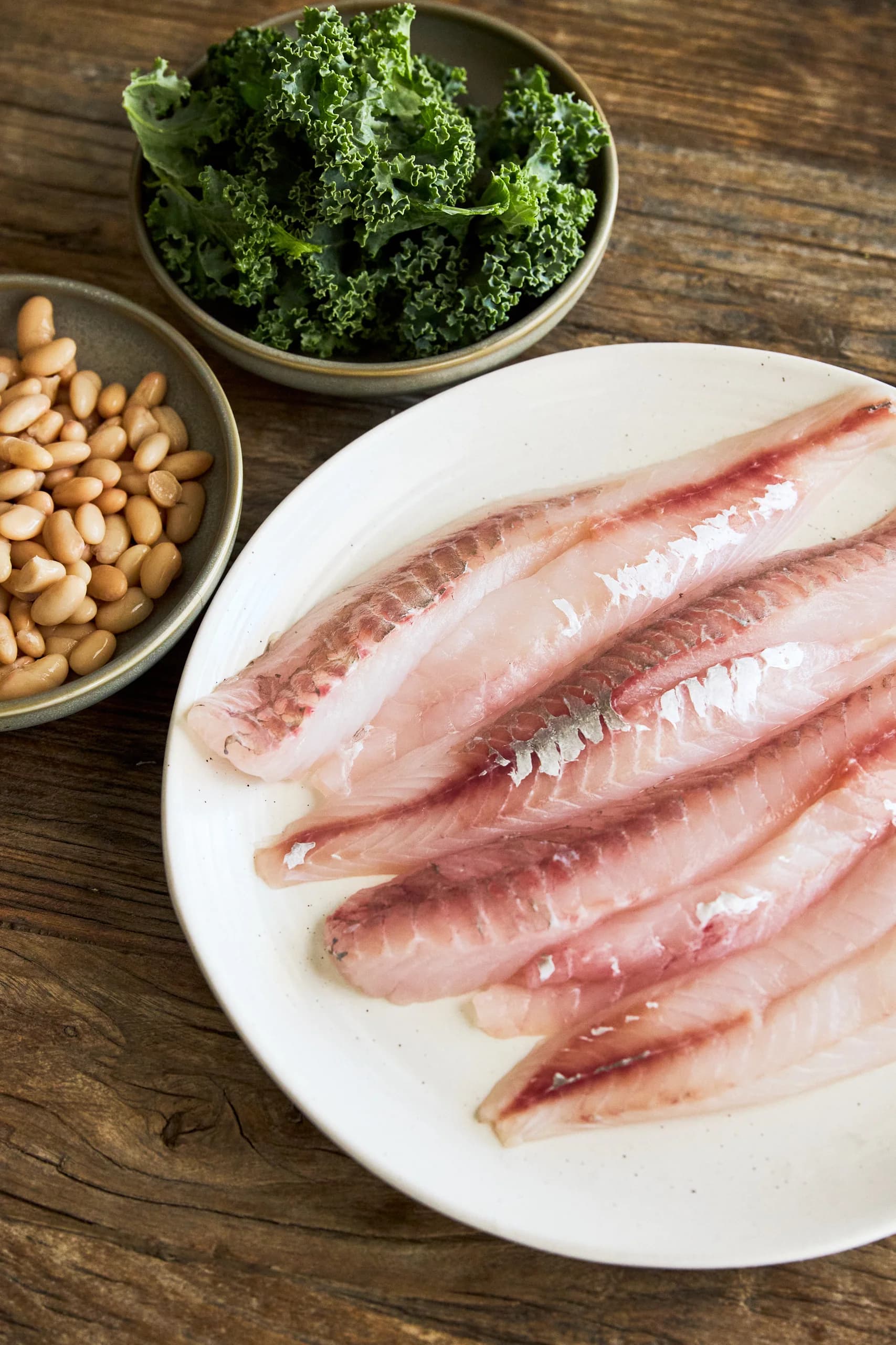 Raw white fish fillets on a cream platter beside bowls of curly kale and white beans on a rustic timber table