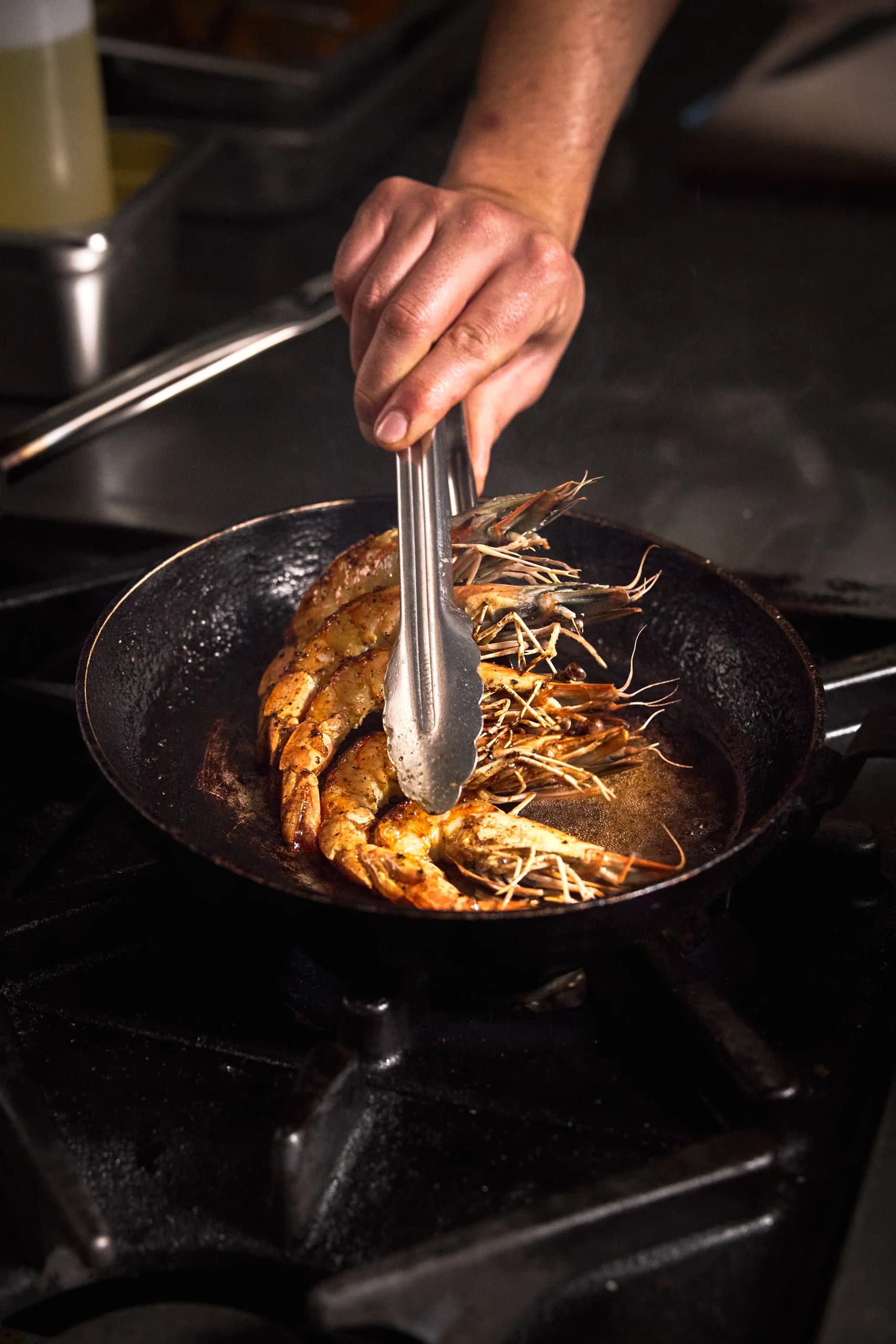 Tongs turning golden seared whole prawns in a black pan over a gas burner