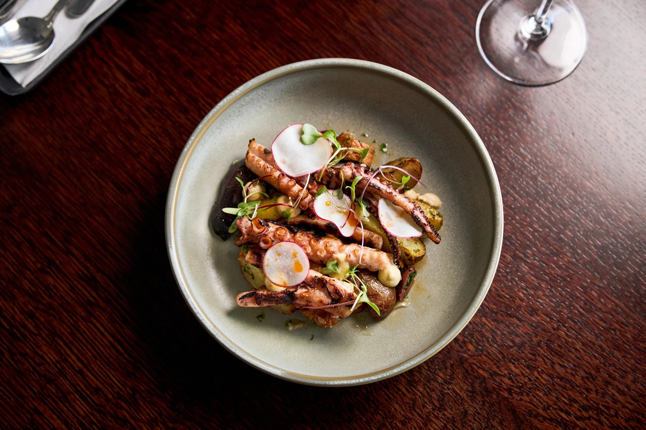 Overhead view of grilled octopus tentacles with roasted potatoes, shaved radish and microgreens in a grey bowl