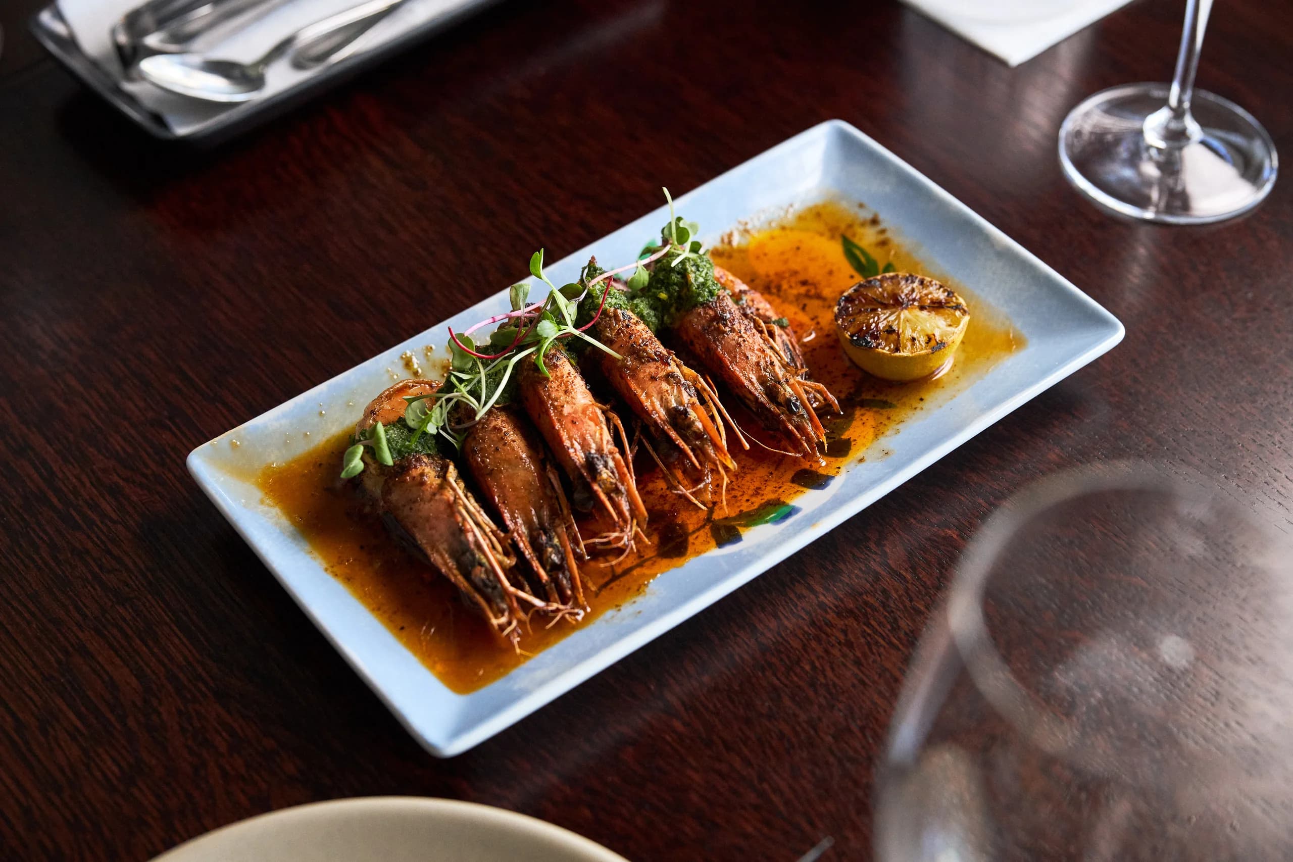 Grilled whole prawns in orange oil with herb sauce and charred lemon on a pale blue plate