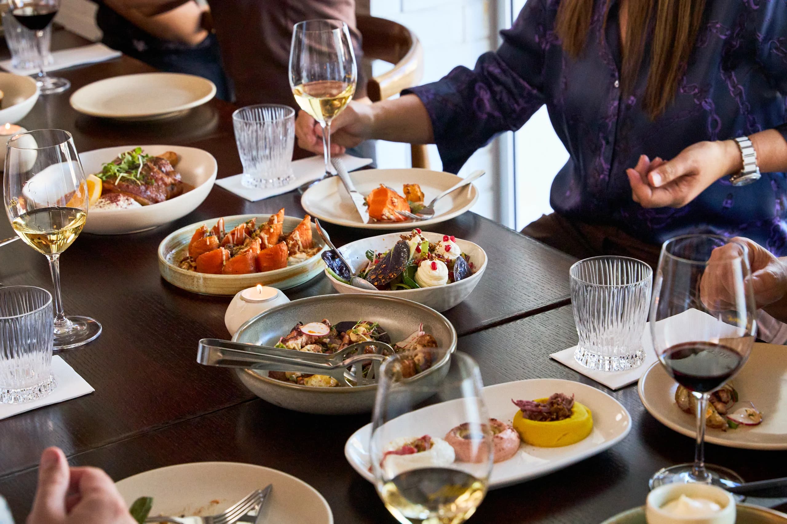Shared plates of roasted carrots, salad, dips and lamb on a dark table with wine and a tealight
