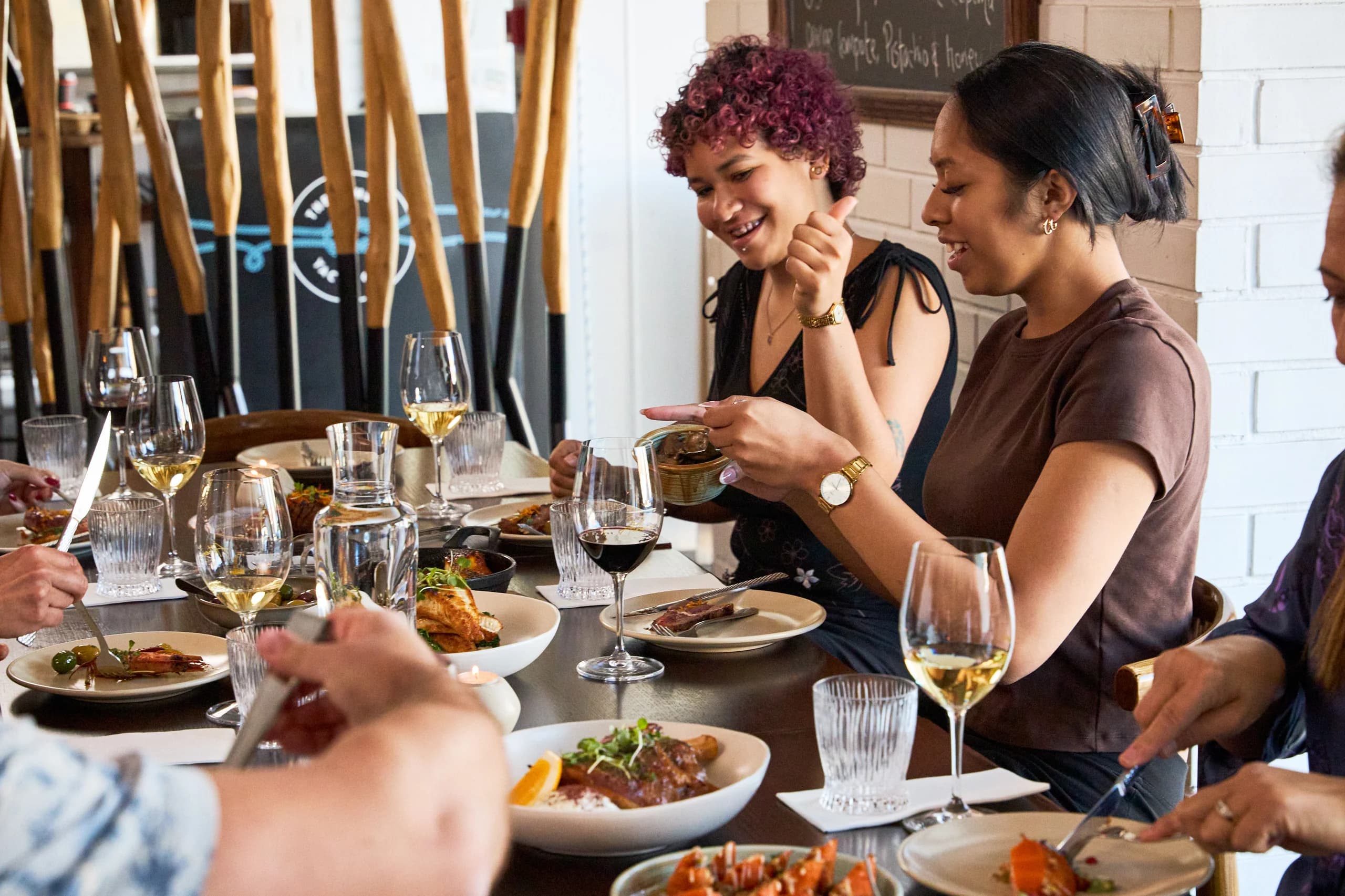 Two diners laughing and sharing a dish at a long table, timber pole screen behind