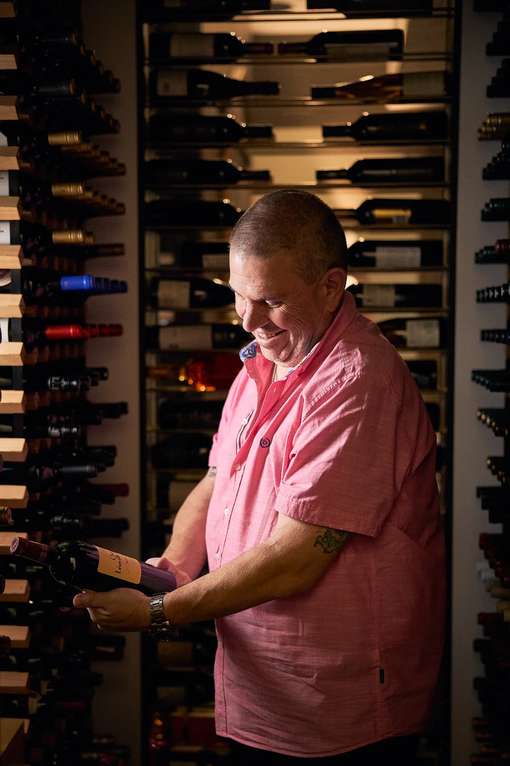 Emilios in the cellar, smiling as he reads the label on a bottle of red surrounded by racks of wine