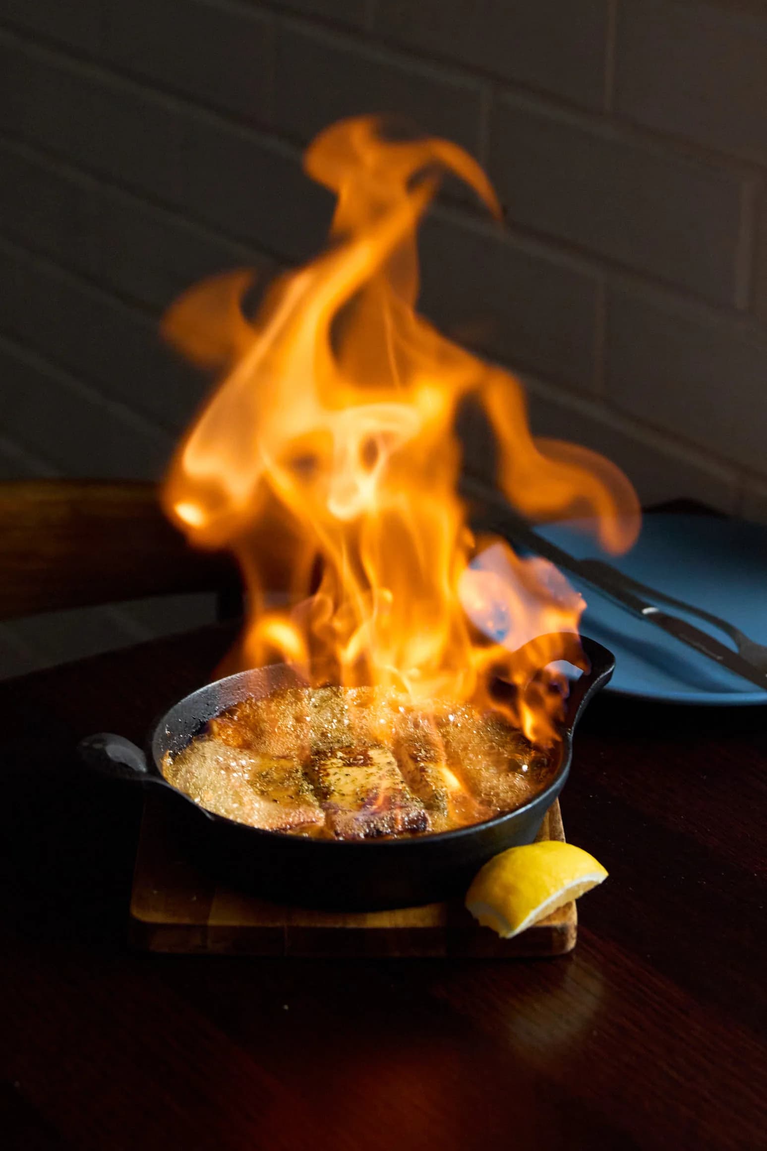 The AYC Flamed Cheese alight in a cast-iron pan, flames rising high above it, with a wedge of lemon on the board