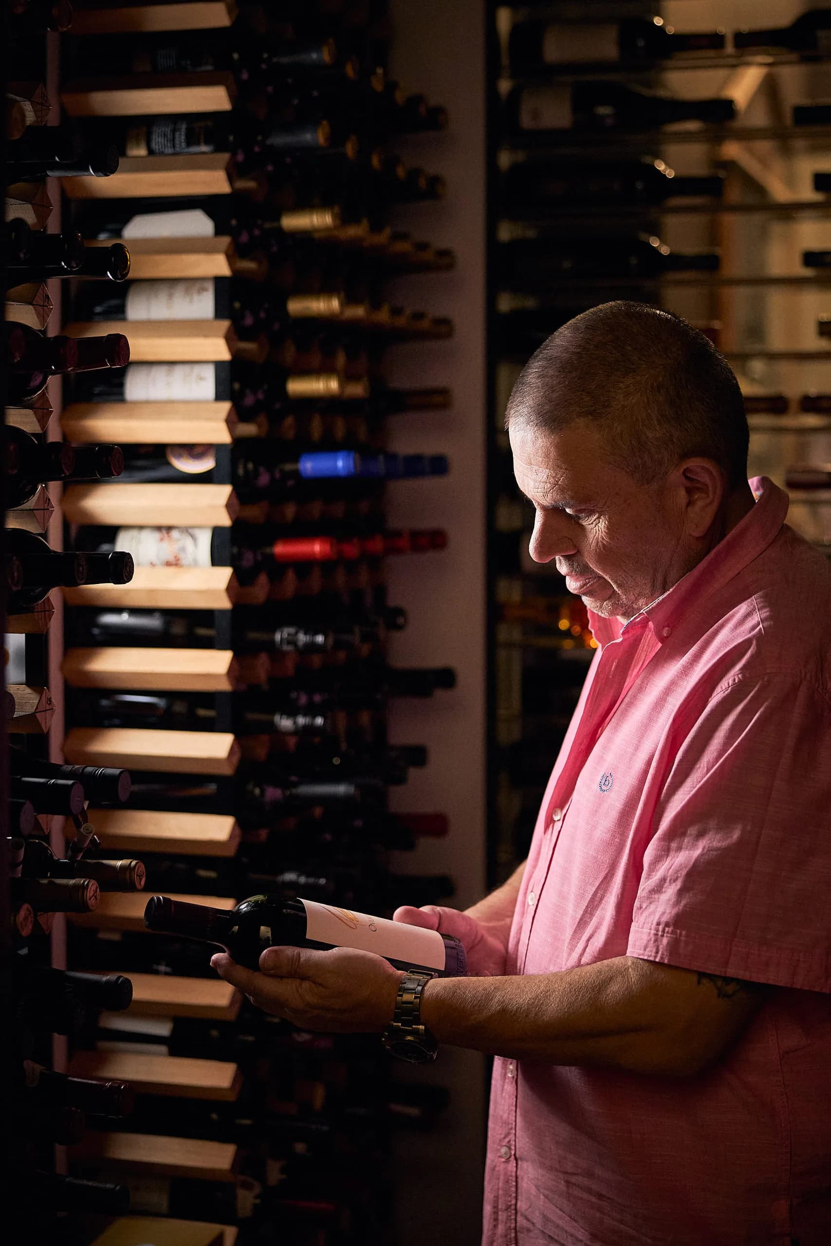 Emilios in the cellar, studying the label on a bottle of red beside a wall of racked wine
